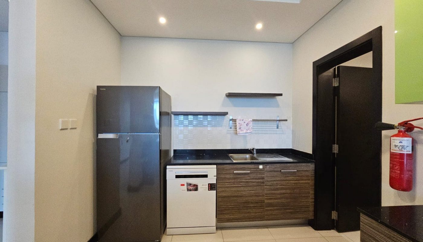A modern flat for rent in Reef Island boasts a kitchen with a stainless steel refrigerator, a dishwasher, a sink, wooden cabinets, and shelves on a tiled backsplash wall. A fire extinguisher is mounted on the right wall.