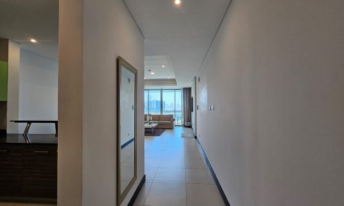 A hallway in a modern flat for rent on Reef Island features white walls and tiled flooring, leading to a living room with large windows showcasing a city view. A large mirror is mounted on one side of the hallway.