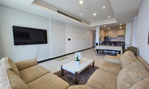 A modern living room in a rental property with a beige sectional sofa, white coffee table with a flower arrangement, flat-screen TV mounted on the wall, and a dining area with table and chairs in the background. Ideal for those seeking a stylish flat for rent near Reef Island.