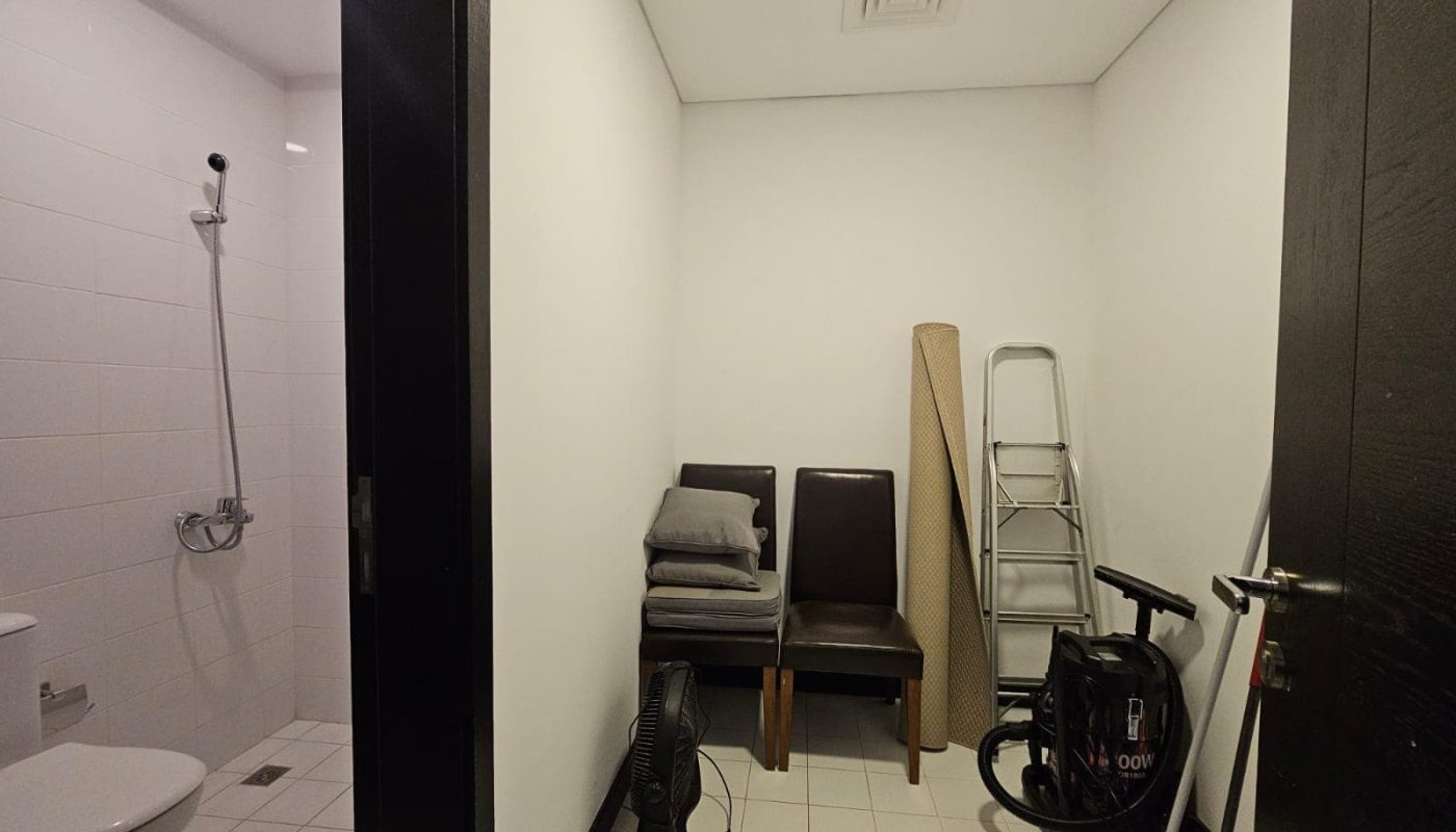 A cozy flat for rent on Reef Island features a small room with cleaning equipment, including a vacuum, fan, ladder, two chairs with folded towels, and a mop. Additionally, the adjacent bathroom is equipped with a toilet, shower, and tiled walls.