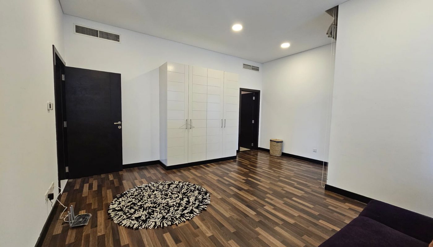 A minimalist flat for rent on Reef Island featuring wooden flooring, white walls, a white wardrobe, black doors, a round patterned rug, and a purple sofa in the corner.