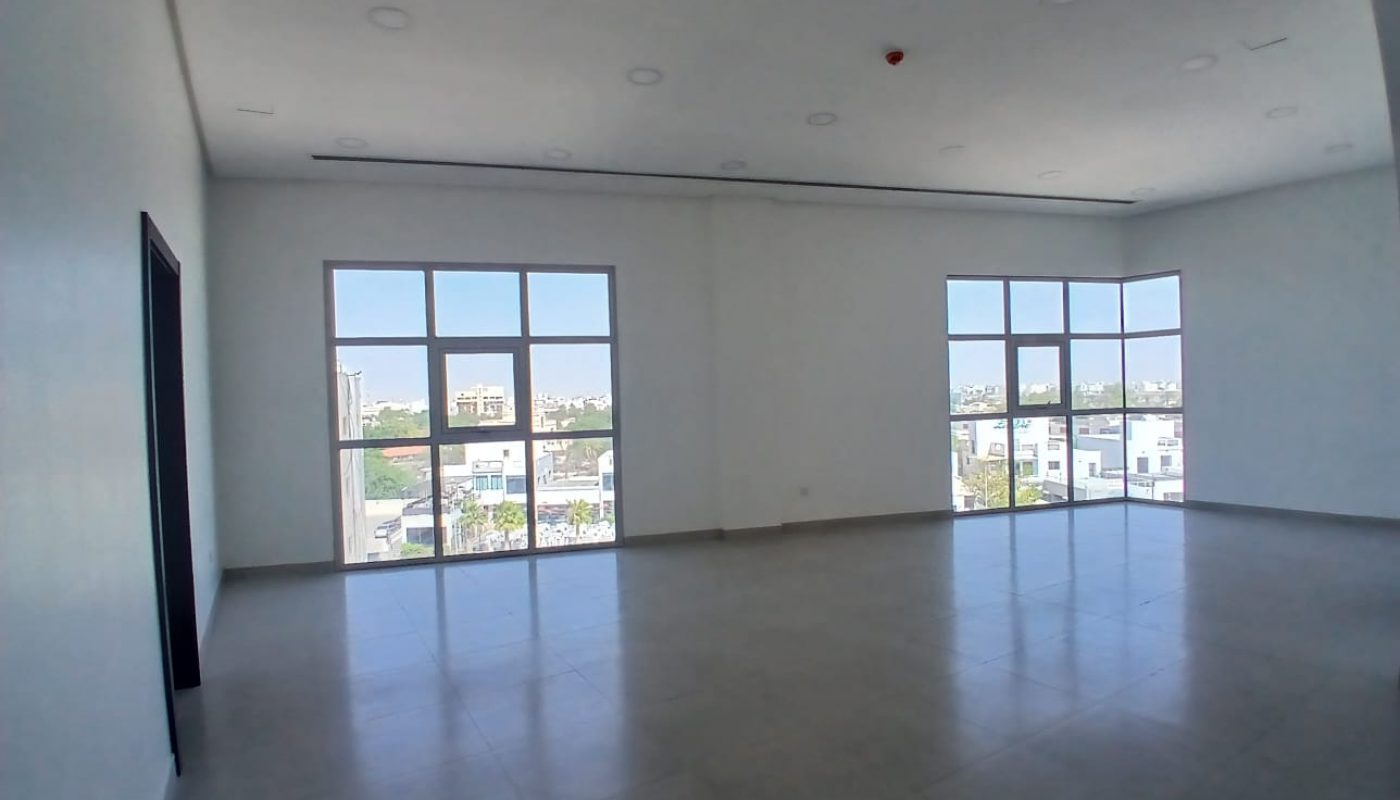 Empty, unfurnished room with large windows on two sides, white walls, tiled floor, and ceiling lights, allowing natural light to enter and offering a view of buildings outside.