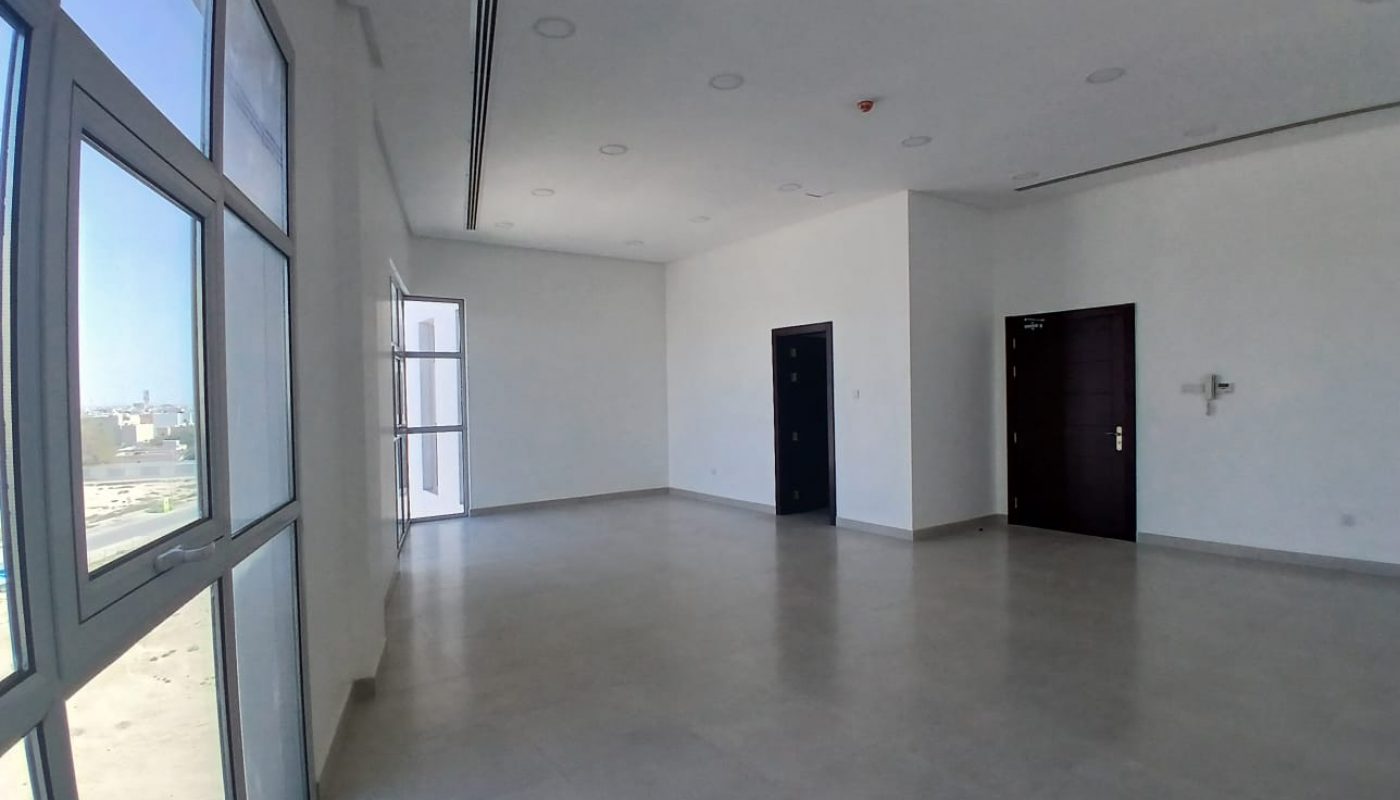 An empty, unfurnished room with white walls, large windows, tiled floor, and two dark doors. Bright natural light enters from the windows on the left.