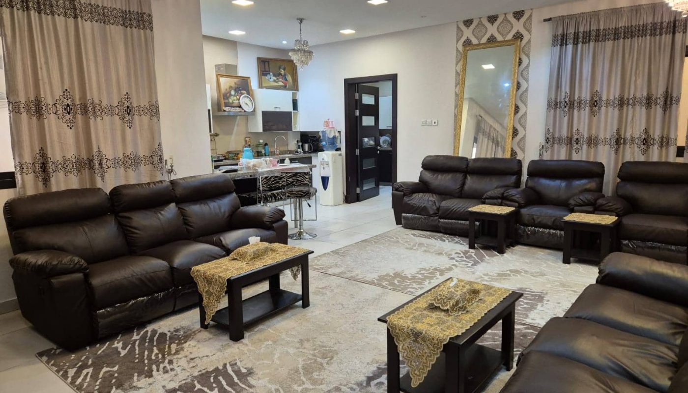 A living room with dark leather sofas, small coffee tables with lace coverings, and patterned curtains. A carpet covers the floor, and a kitchen is visible in the background.