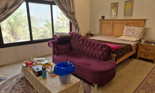 A bedroom with a bed, a purple sofa, a coffee table with various items, and a large window with a view of trees.