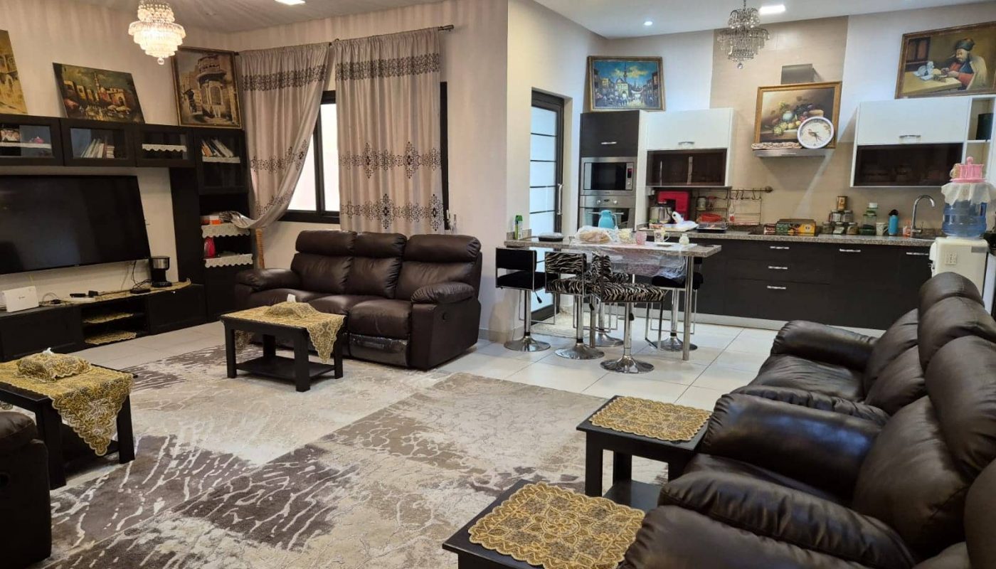 A modern living room and kitchen in a villa for sale in Jid Ali, featuring brown leather sofas, a large TV, a kitchen island with bar stools, chandeliers, and wall art. The space boasts patterned rugs and lace-trimmed table covers.