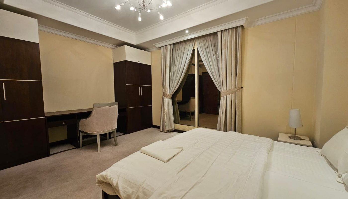 This Juffair flat for rent features a cozy bedroom with a bed, white linens, a bedside table, a desk with a chair, and a modern light fixture. Large closets and sliding doors with beige curtains lead to a balcony, making it an ideal rental.