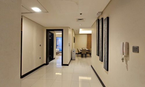 A hallway with beige walls and white tile flooring, leading to an open dining area and a bedroom. A wall-mounted phone and framed art are visible on the right wall.