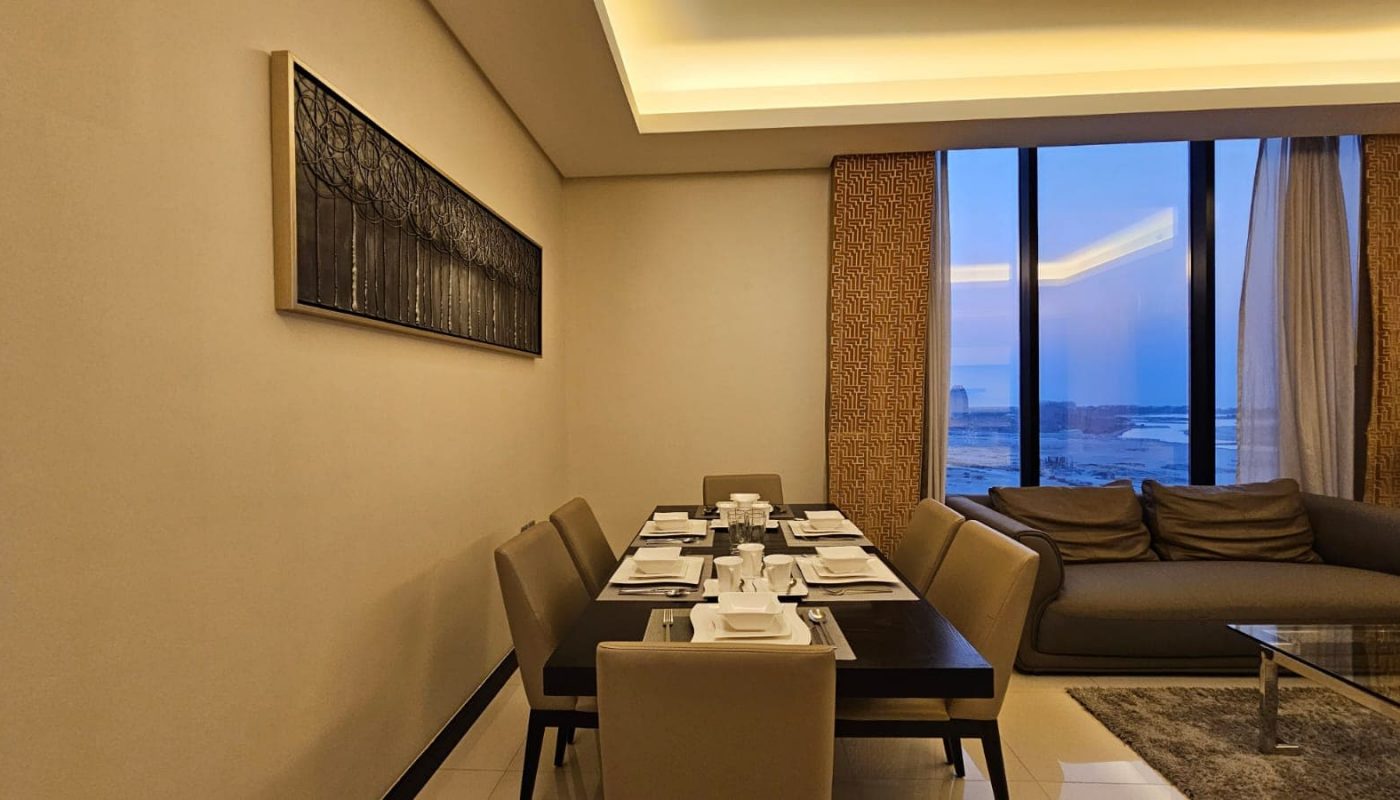 A modern dining room with a table set for eight, beige chairs, a couch, a large window with curtains, and a piece of wall art.