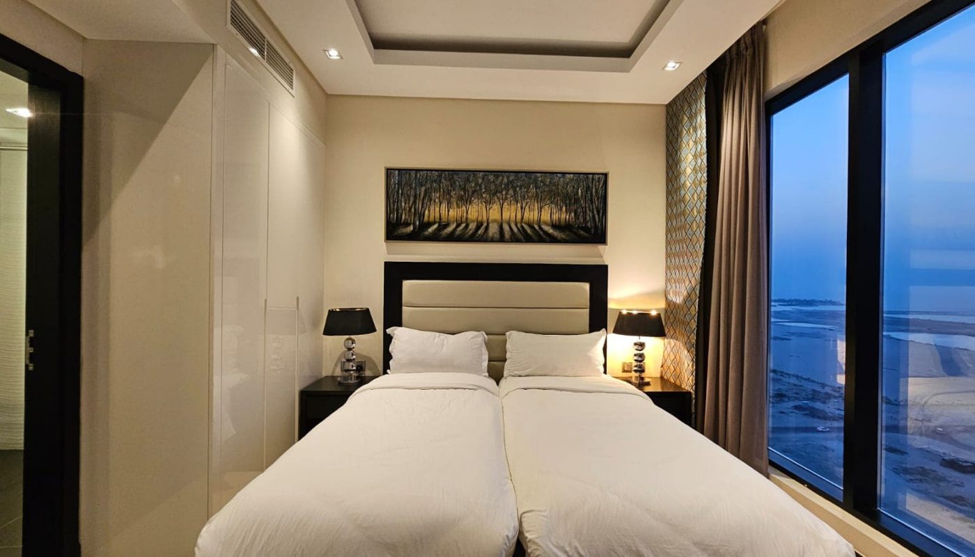 A modern bedroom features two single beds with white bedding, bedside tables with lamps, a window with a coastal view, and a framed artwork of trees on the wall above the beds.