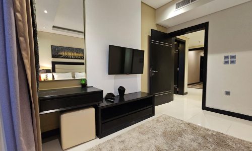 A modern hotel room with a wall-mounted TV, a black dresser, a mirror reflecting a bed, an ottoman, and open doorways leading to other rooms. The floor is white tiled with a beige rug.