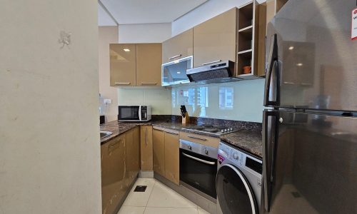 Modern kitchen in a Juffair flat for rent with beige cabinets, granite countertops, and stainless steel appliances including a refrigerator, oven, microwave, and washing machine. A small sink is visible on the left.