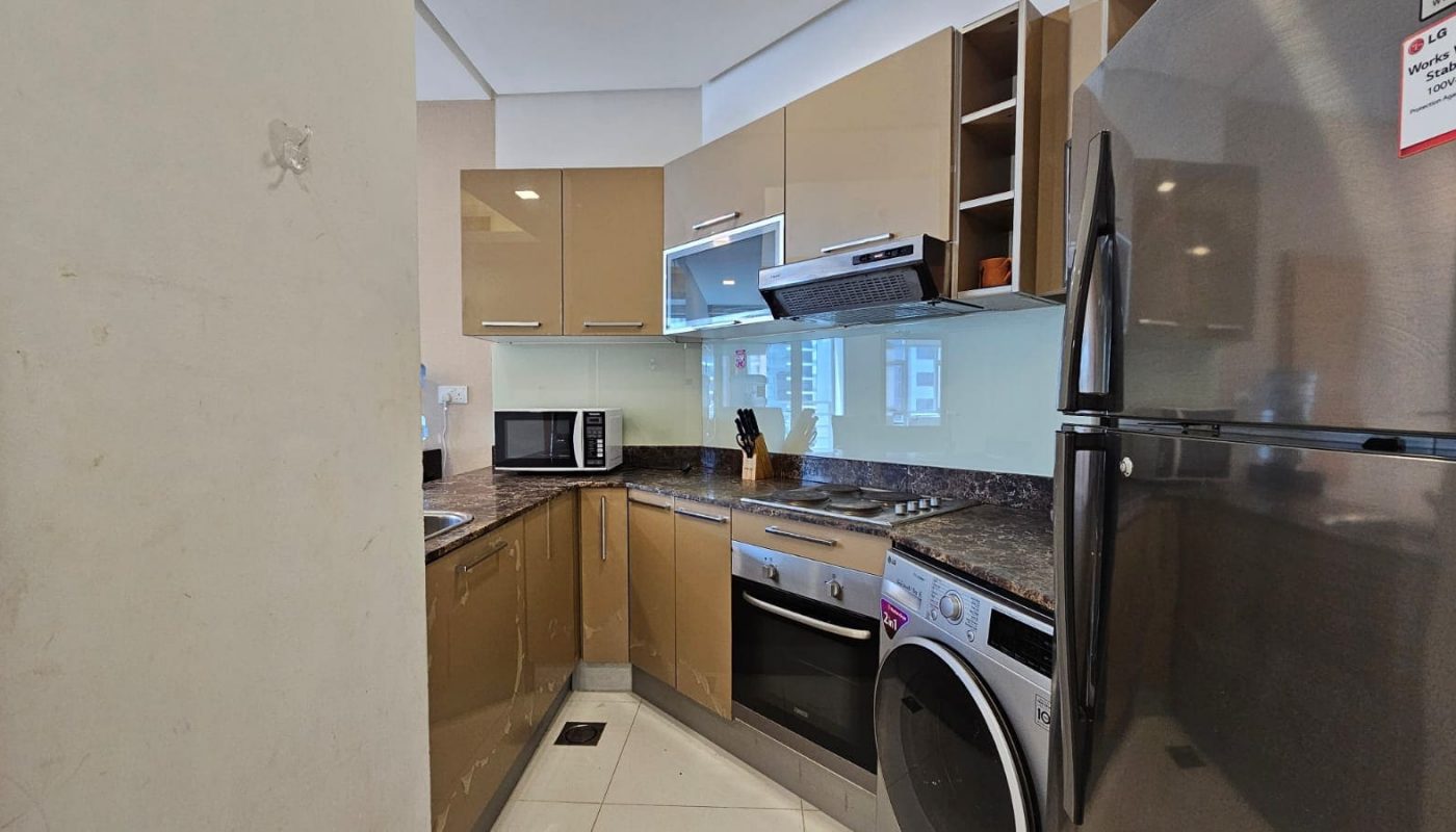 Modern kitchen in a Juffair flat for rent with beige cabinets, granite countertops, and stainless steel appliances including a refrigerator, oven, microwave, and washing machine. A small sink is visible on the left.
