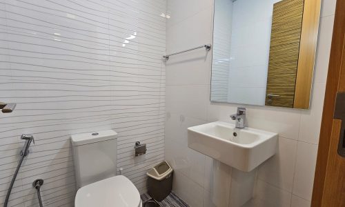 A clean bathroom in a Juffair apartment rental features a white toilet, sink, mirror, and beige trash bin. White tiles with gray lines cover the wall, and a wooden door is partially visible.