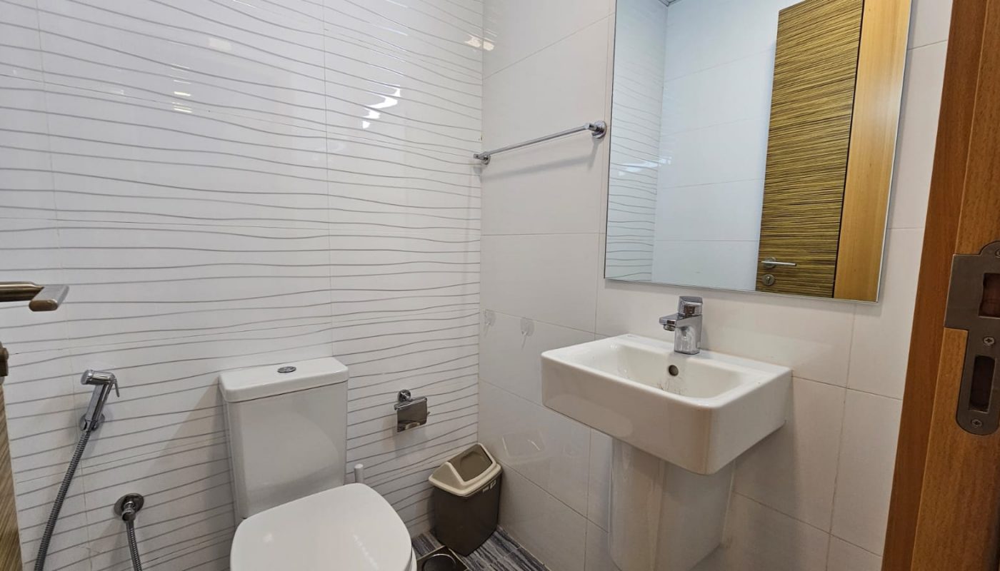 A clean bathroom in a Juffair apartment rental features a white toilet, sink, mirror, and beige trash bin. White tiles with gray lines cover the wall, and a wooden door is partially visible.