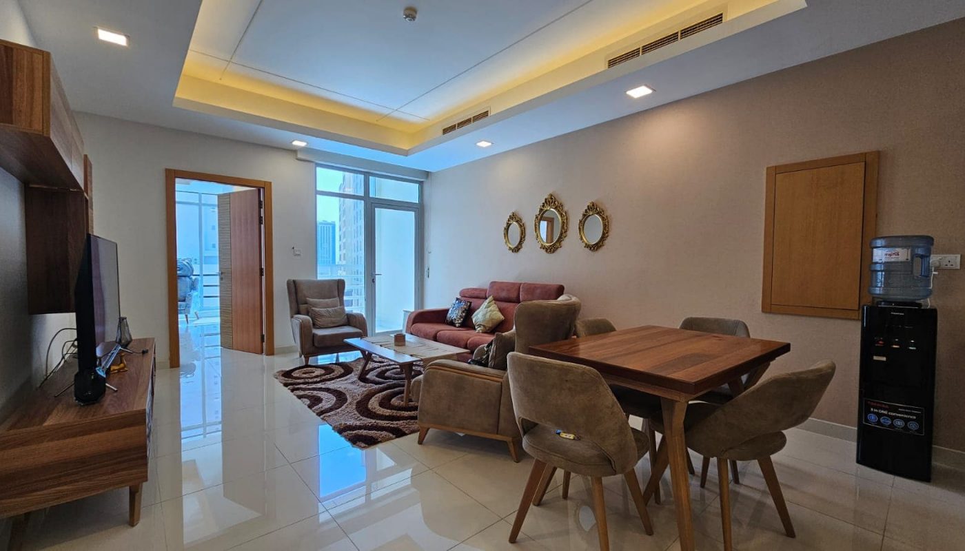 Discover a stylish flat for rent in Juffair featuring a modern living and dining area with a wall-mounted TV, brown sofa, armchairs, wooden dining table with chairs, decorative mirrors, and a door leading to a balcony.
