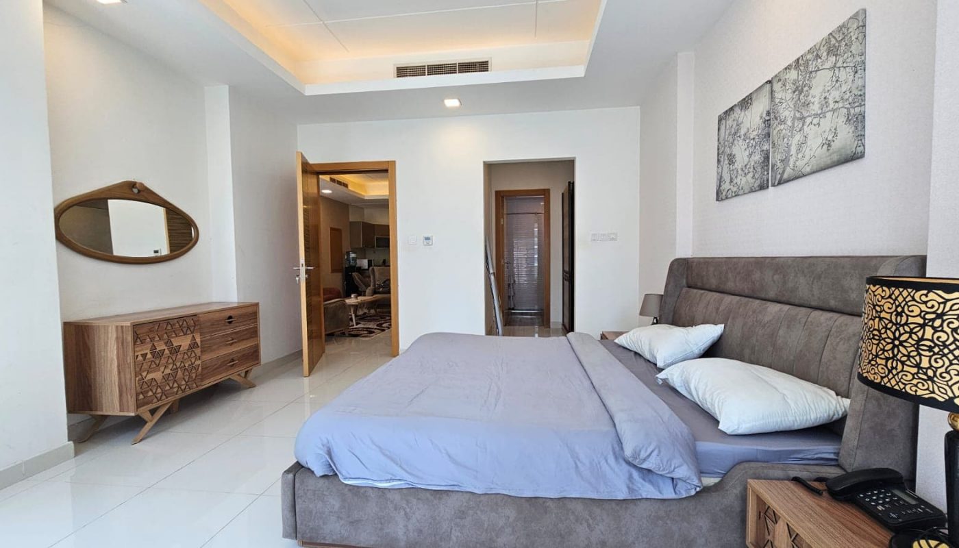 Modern bedroom in a Juffair flat for rent featuring a gray upholstered bed, two pillows, a nightstand with a lamp, a wooden dresser, and a mirror on the wall. The room has white walls, tiled floors, and an open door leading to a hallway.
