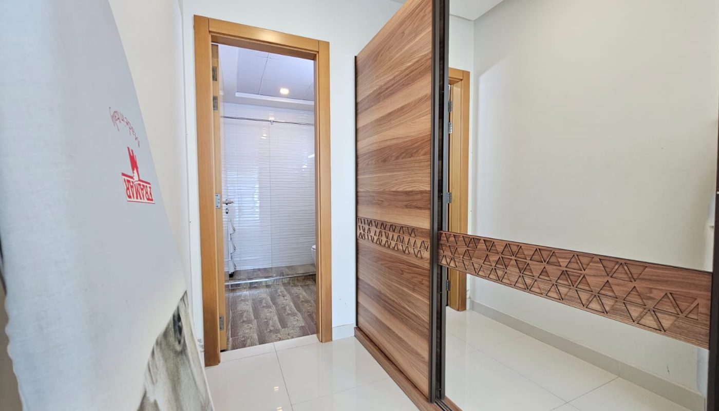 A hallway in a rental property with a large mirror on the right and a wooden door at the end leading to a bathroom with a glass shower. There is a mattress standing against the left wall. This charming flat for rent is located in the desirable Juffair area.