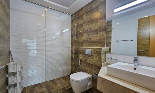 Modern bathroom in a Juffair flat for rent features a glass-enclosed shower, wall-mounted toilet, rectangular sink on a beige countertop, and wooden accent walls. A mirrored cabinet is above the sink, and a wooden door is visible.