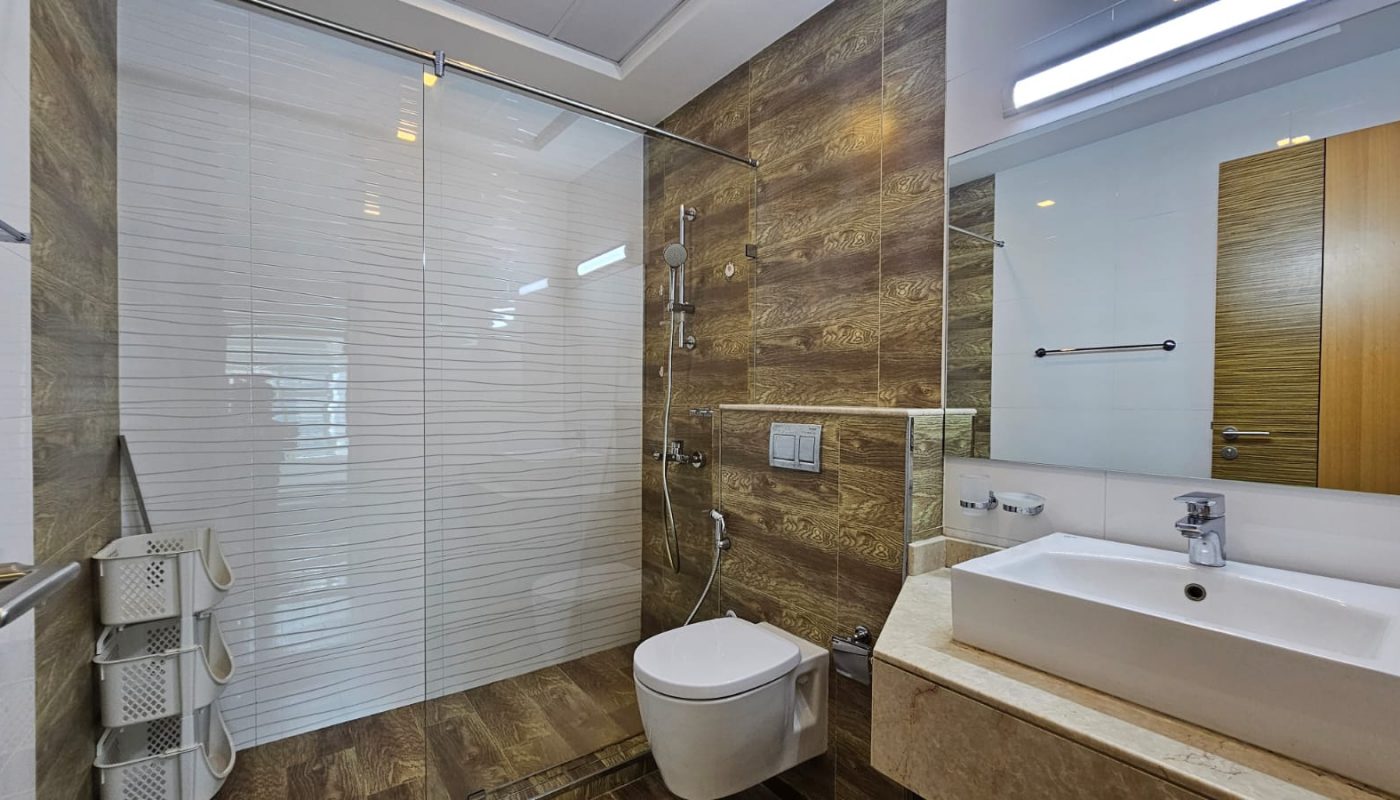 Modern bathroom in a Juffair flat for rent features a glass-enclosed shower, wall-mounted toilet, rectangular sink on a beige countertop, and wooden accent walls. A mirrored cabinet is above the sink, and a wooden door is visible.