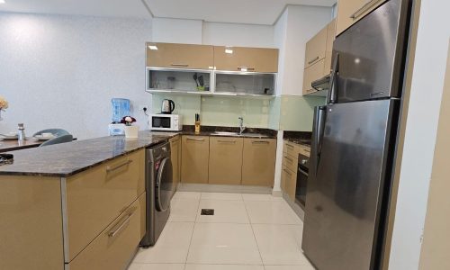 Modern kitchen in a luxury apartment with beige cabinets, stainless steel appliances including a refrigerator, oven, microwave, and washing machine. Countertop items include a water dispenser, toaster, and kettle. Ideal for those seeking upscale living in a Juffair rental.