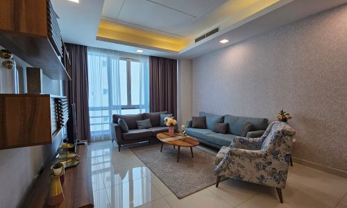 A modern luxury apartment living room in Juffair with a large window, two sofas, a patterned armchair, a wooden coffee table on a rug, and a wall-mounted bookshelf.