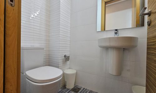 A small bathroom in a modern apartment with a white toilet, round sink, wall mirror, and trash bin. The walls feature pristine white tiles, and the floor boasts sleek gray tiles. A wooden door is partially visible on the right, completing this chic Juffair rental space.