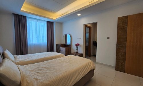 A modern bedroom in a luxury apartment featuring two single beds, a dresser with a mirror, a wooden door, and an ensuite bathroom. The room boasts a window with white curtains and recessed ceiling lighting.