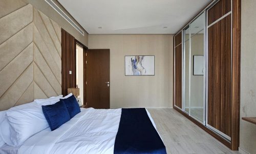 A modern bedroom with a bed featuring white and navy bedding, a wooden door, a wall-mounted abstract painting, and mirrored sliding closet doors along the right wall.