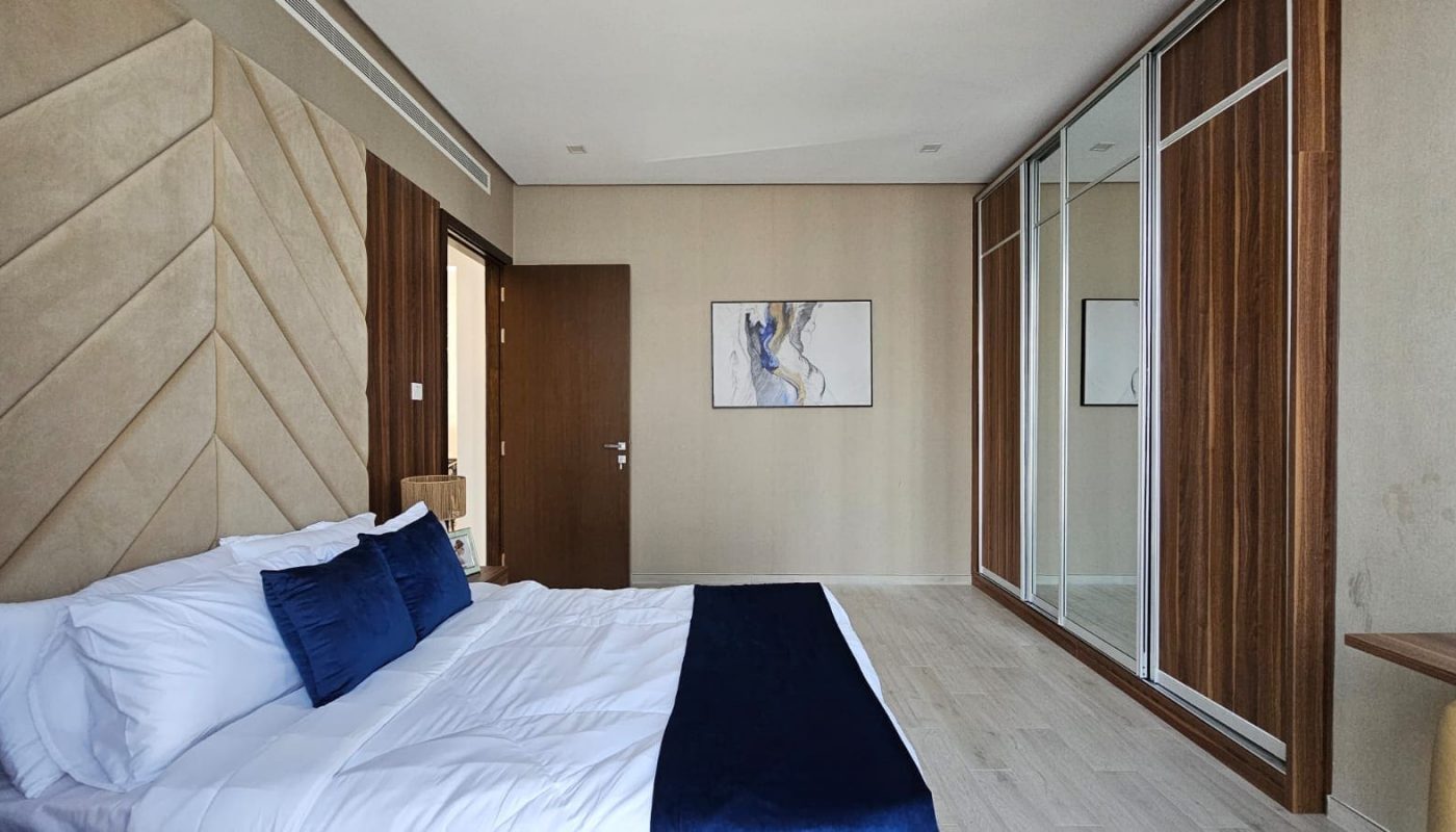 A modern bedroom with a bed featuring white and navy bedding, a wooden door, a wall-mounted abstract painting, and mirrored sliding closet doors along the right wall.