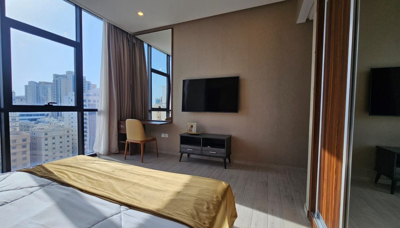 Modern bedroom with a large window, city view, a wall-mounted TV, a desk with a chair, and a bed with a yellow blanket. There is a photograph on the desk and a closet with mirrored doors.