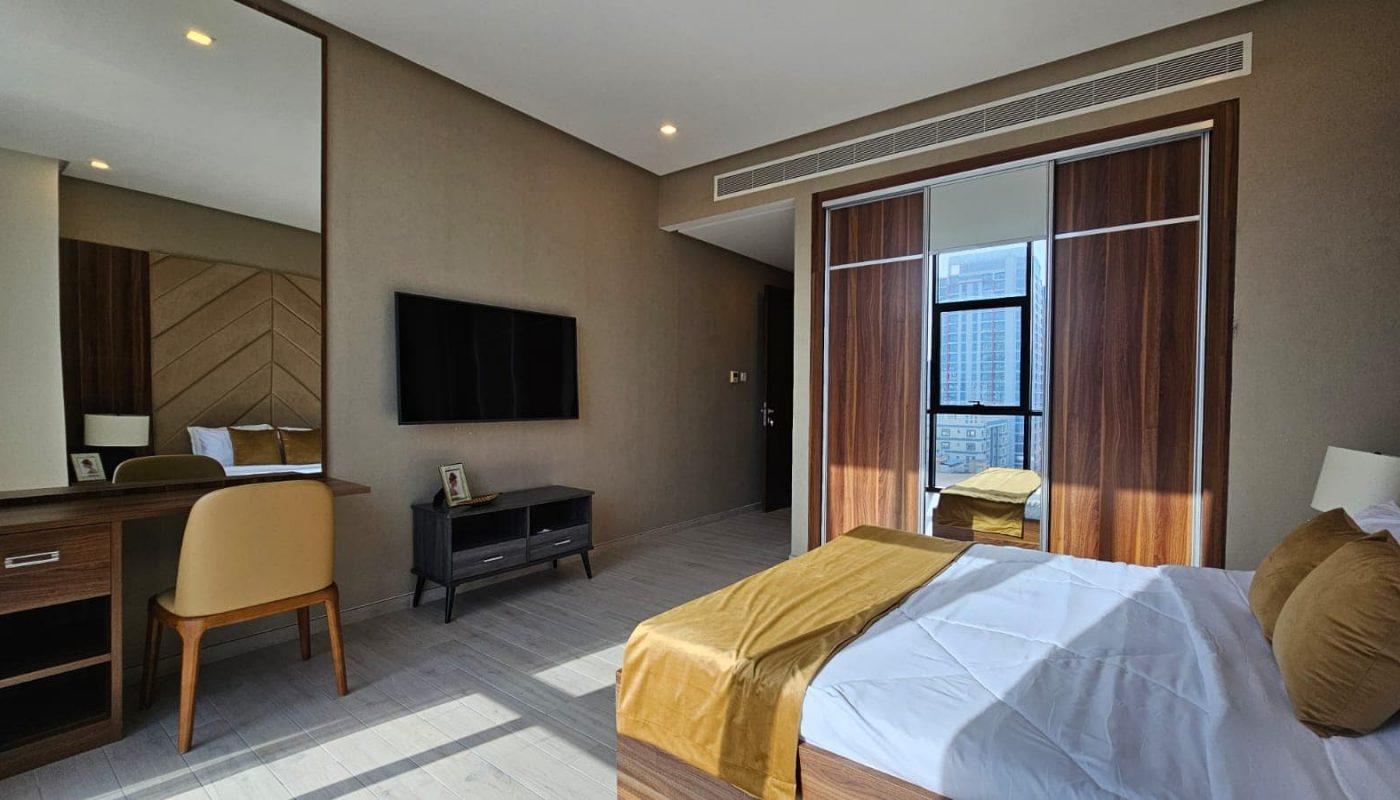 A contemporary hotel room with a bed, desk, wall-mounted TV, and large window. The decor is modern, featuring wooden furniture and a neutral color palette with gold accents.