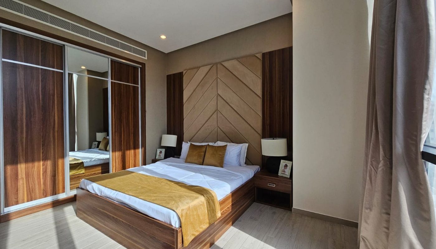 A modern bedroom with a large bed featuring brown and beige bedding, wooden accents, mirrored sliding door closet, two bedside tables with lamps, and a large window with beige curtains.