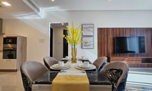 A modern dining table set with yellow flowers in the center, surrounded by upholstered chairs. In the background, there’s a wall-mounted TV and contemporary decor.