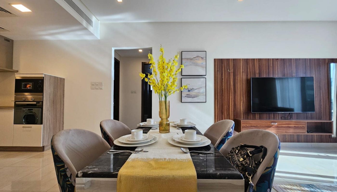 A modern dining table set with yellow flowers in the center, surrounded by upholstered chairs. In the background, there’s a wall-mounted TV and contemporary decor.