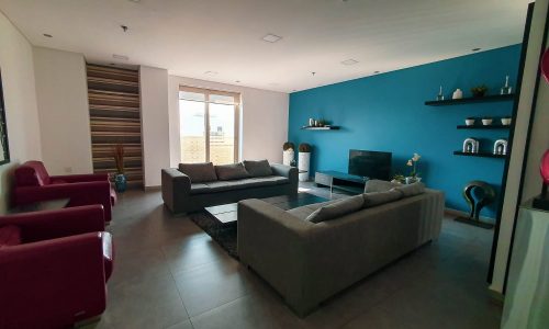 This stylish flat for rent in Juffair features a modern living room with a blue accent wall and two gray sofas facing a TV. Red armchairs, wall-mounted shelves, and a large window complete the inviting space. Perfect for anyone seeking an elegant rental property.