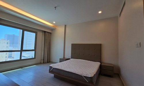 A well-lit bedroom in a Juffair flat for rent, featuring a large window, a double bed with a tufted headboard, and two nightstands. The room has light-colored walls and a tiled floor, with city buildings visible through the window.