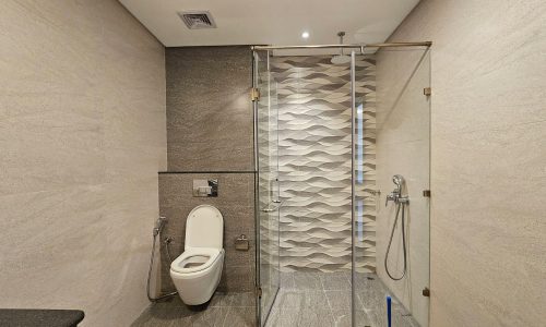 Modern bathroom in a flat for rent in Juffair, featuring a toilet, glass-enclosed shower, and decorative wave-patterned tiles on the shower wall. The room boasts light-colored walls and floor tiles with a sleek, minimalistic design, perfect for a rental property.