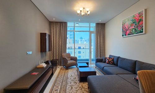 A modern living room in a Juffair flat for rent features a gray L-shaped couch, an armchair, a coffee table, a mounted TV unit, a decorative rug, and large windows with curtains. A framed painting adds an artistic touch to the space.