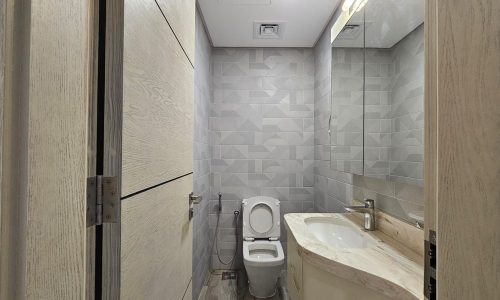 A modern bathroom in a flat for rent in Juffair features light gray geometric tile walls, a toilet, a sink with a marble countertop, and a large mirror above the sink. There is an open door on the left.