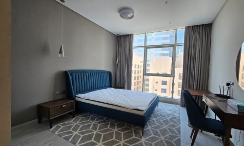 A modern, minimalist bedroom in a Juffair flat for rent features a blue upholstered bed, a bedside table, and a wooden desk and chair set. Large windows with gray curtains provide stunning views of the urban buildings outside. This stylish rental property offers both comfort and convenience.