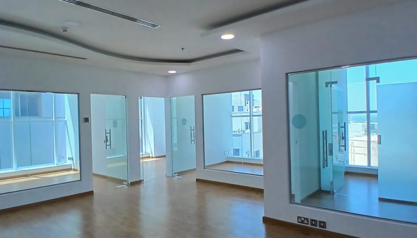 A spacious, modern office in Seef District with wooden flooring, ceiling lights, and floor-to-ceiling glass walls dividing the space into separate rooms. This fitted office offers a sea view that adds to its appeal.