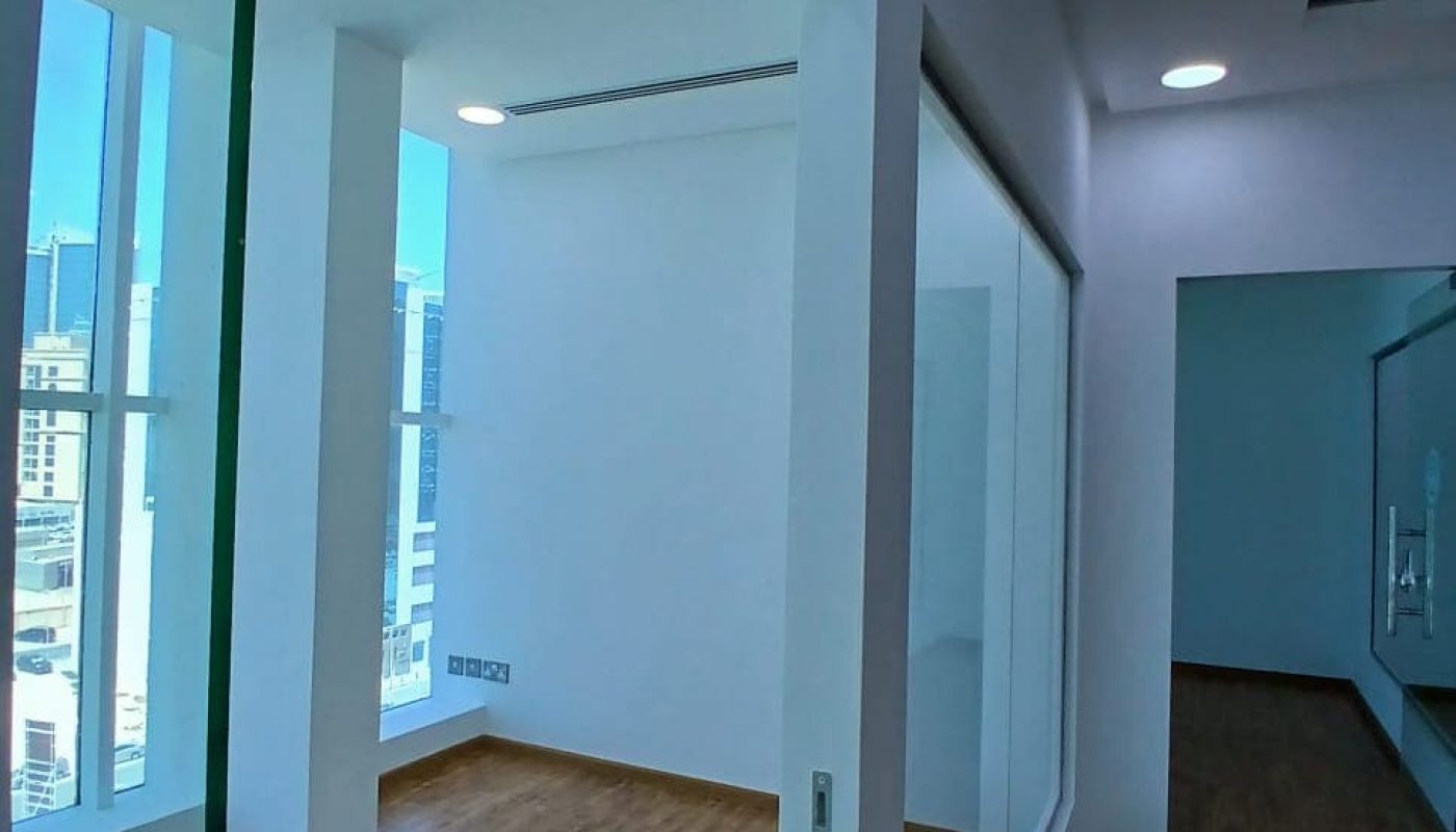 Empty office space with glass walls, wooden flooring, and a partial view of urban buildings in the Seef District outside the windows.