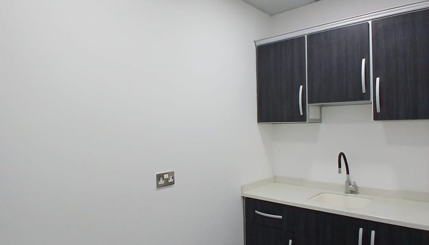 A small corner kitchen with dark wooden cabinets, a beige countertop, a single sink, and overhead lighting in a white-walled room in the Seef District.