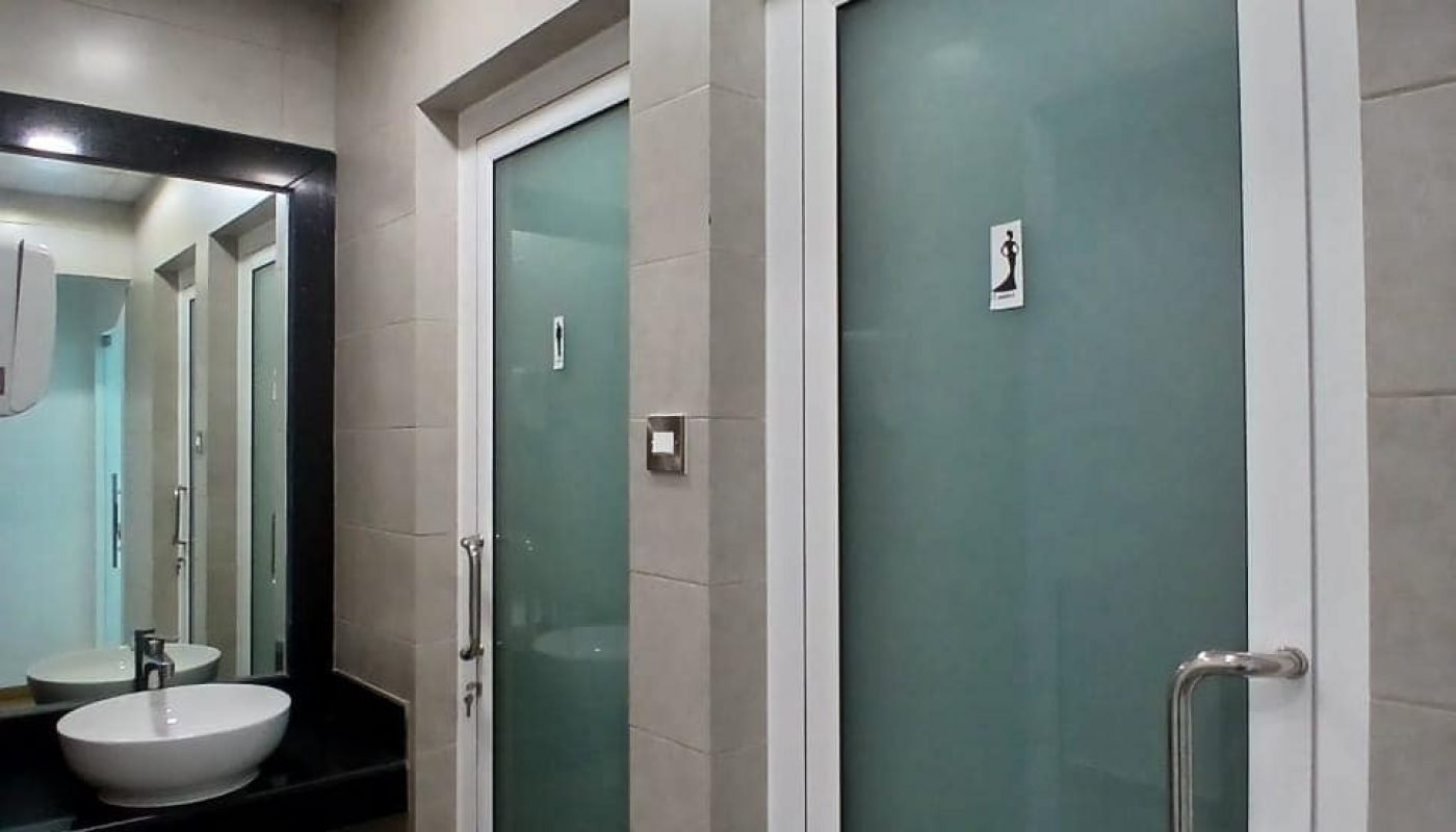 A bathroom in the Seef District with two closed doors featuring frosted glass, each marked with a gender sign, a sink with a mirror, soap dispenser, and overhead lighting.