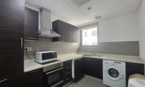 A modern kitchen in a Juffair flat for rent, featuring dark wood cabinets, stainless steel appliances, a microwave on the counter, a washing machine, and a window above the sink.