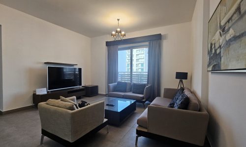 A modern living room in a Juffair apartment rental features a sofa, armchair, and a glass coffee table facing a large flat-screen TV. There's a window with curtains and blinds, and a painting on the wall. The room has tile flooring. This stylish flat for rent offers comfort and convenience in every corner.