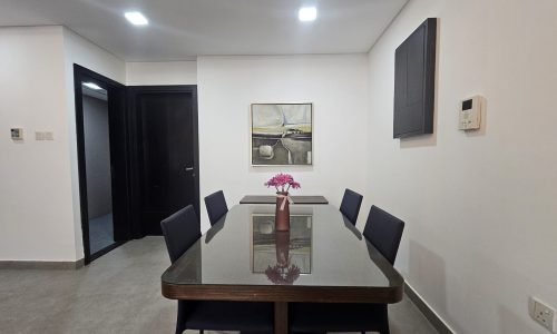 A stylish Juffair flat for rent featuring a glass-top dining table with six chairs, a vase with pink flowers centered on the table, and a large painting on the wall. Two doors in the background complete this elegant rental property setting.