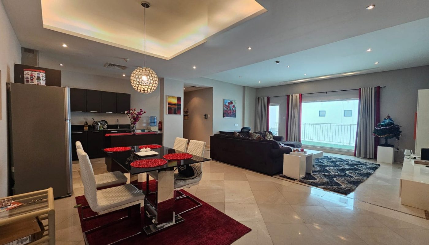 Modern living room and dining area with an open floor plan. The space includes a dining table, kitchen, couches, TV, large window with balcony, and various decorations.