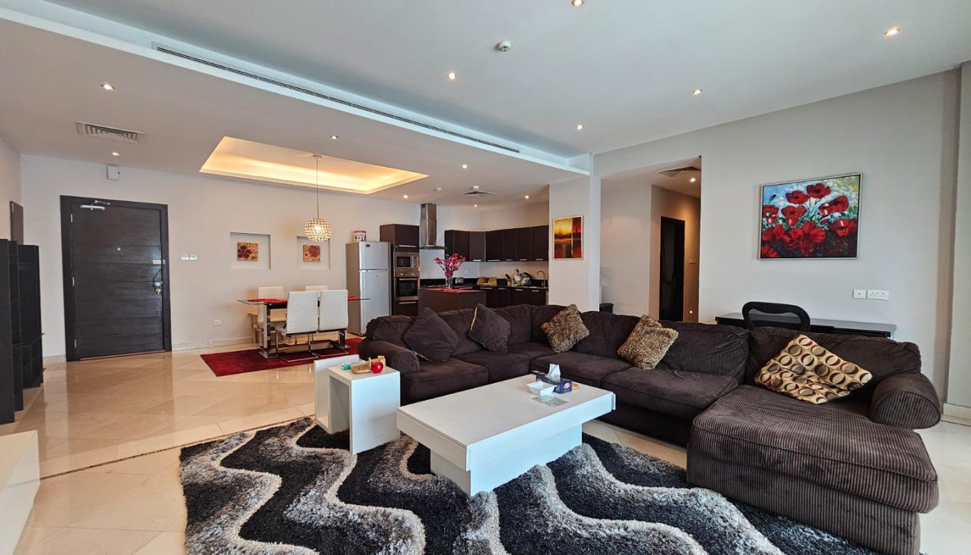 A modern living room with a dark sectional sofa, a white coffee table, and a patterned rug. The room extends into an open-plan kitchen and dining area, featuring a table with white chairs.